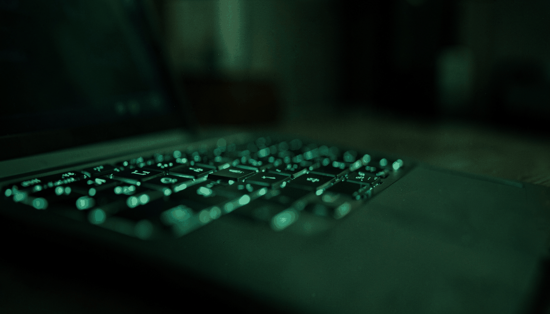A softly lit close-up of a modern laptop keyboard with a subtle green-tinted glow reflecting off the keys, set against a dark blurred background suggesting a calm home-office environment.