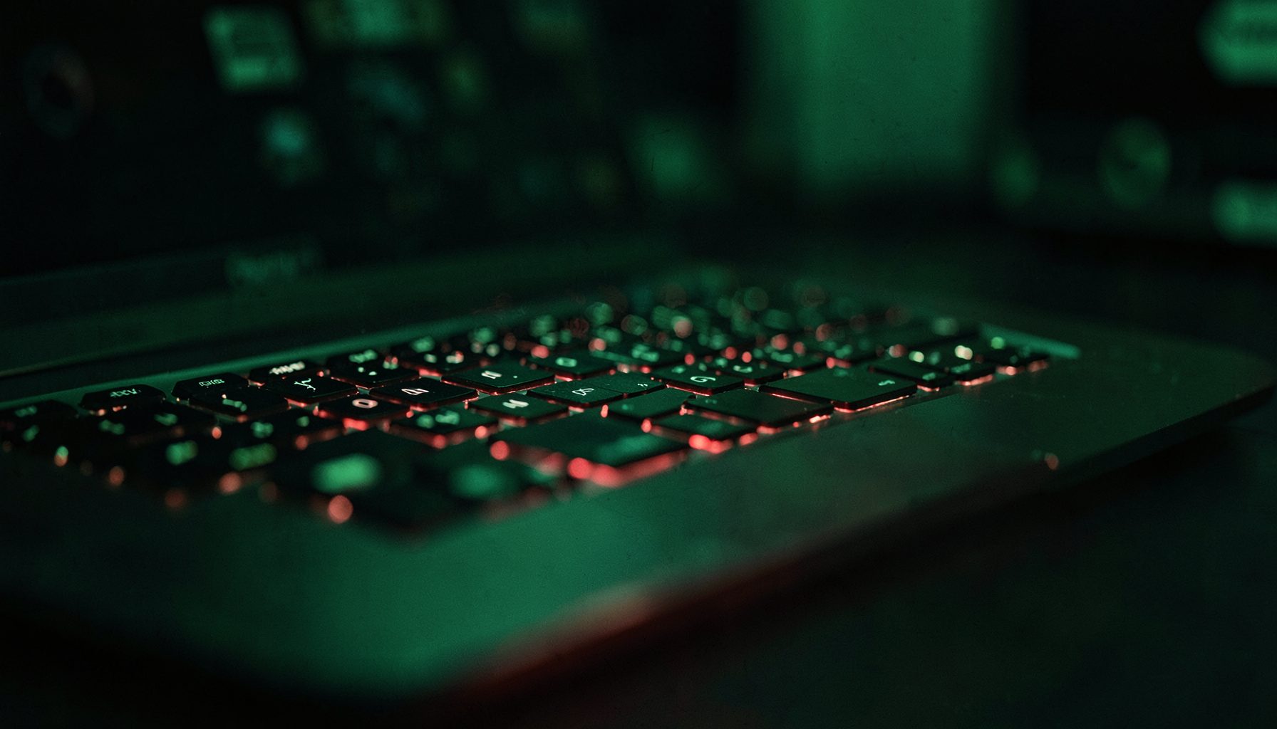 A wide shot of a laptop keyboard with a subtle red glow emanating from beneath the keys, shallow depth of field, dark and moody cybersecurity theme